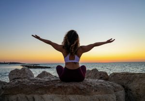 A woman sits on rocks at sunset, arms outstretched, embracing the beauty of the moment and the vibrant sky