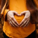 A pregnant woman in an orange shirt and scarf forms a heart shape with her hands, radiating joy and love