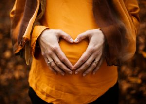 A pregnant woman in an orange shirt and scarf forms a heart shape with her hands, radiating joy and love