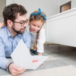 A father and daughter lie on the floor together, reading a letter with smiles on their faces