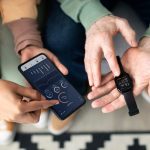 A man and woman are smiling while holding their smart watches, showcasing the devices on their wrists